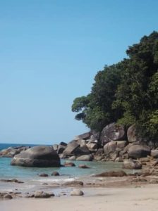 Strand Meer mit großen Steinen, rechts grüne Bäume blauer Himmel